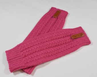 May include: A pair of bright pink knit fingerless gloves featuring a textured cable knit pattern and ribbed cuffs. Each glove is accented with a small, rectangular brown leather tag stitched near the top edge, displaying visible text.
