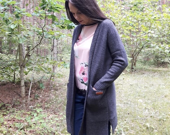 Puede incluir: Una mujer lleva una chaqueta de punto gris con una blusa de estampado floral y unos vaqueros azules. La chaqueta tiene un bolsillo de parche en el lado izquierdo.
