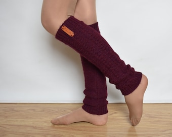 May include: A pair of deep burgundy knit leg warmers featuring a ribbed texture and a small rectangular brown leather tag attached to the upper cuff. The leg warmers extend from the lower calf to the ankle, showcasing a soft, textured yarn construction.