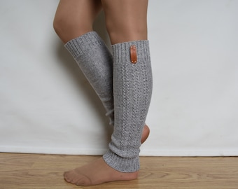 May include: A pair of light gray knit leg warmers featuring a vertical cable knit pattern and a ribbed cuff at the top and bottom. A small, rectangular brown leather tag is attached to the upper ribbed section of the right leg warmer.
