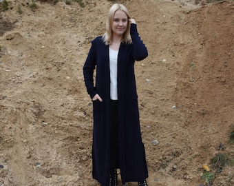 Könnte beinhalten: Eine lange, dunkel marineblaue Strickjacke mit offener Vorderseite, langen Ärmeln und zwei großen aufgesetzten Taschen. Das Kleidungsstück wird über einem schlichten weißen T-Shirt und einer schwarzen Hose getragen, kombiniert mit schwarzen Schnürstiefeln.