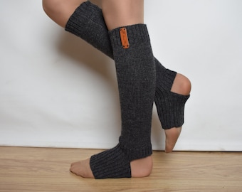 May include: A pair of dark charcoal gray knit leg warmers featuring a ribbed texture and an open heel design. Each leg warmer includes a small, rectangular tan leather-colored brand tag attached near the top cuff, providing a subtle contrast.