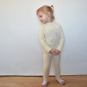 May include: A young child wearing a white knitted sweater and matching pants. The child is standing in front of a white wall.