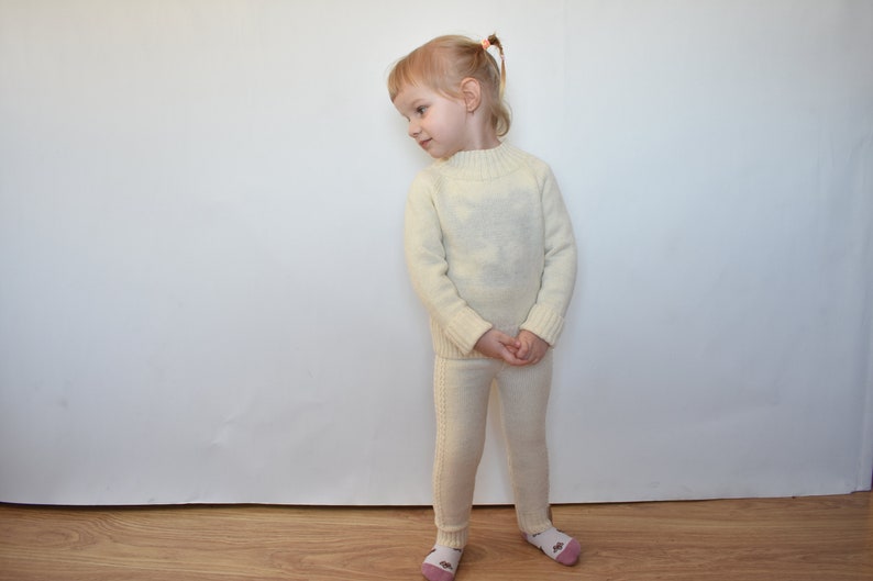 May include: A young child wearing a white knitted sweater and matching pants. The child is standing in front of a white wall.