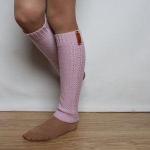 May include: A pair of light pink knit leg warmers featuring a ribbed cuff at the top and bottom, a vertical cable knit pattern along the side, and a small rectangular brown leather-like tag attached to the upper outer edge of the left warmer.