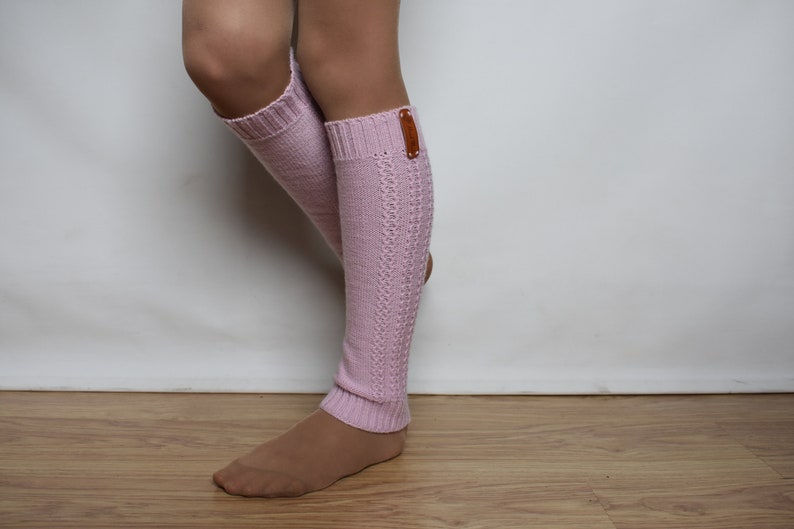 May include: A pair of light pink knit leg warmers featuring a ribbed cuff at the top and bottom, a vertical cable knit pattern along the side, and a small rectangular brown leather-like tag attached to the upper outer edge of the left warmer.