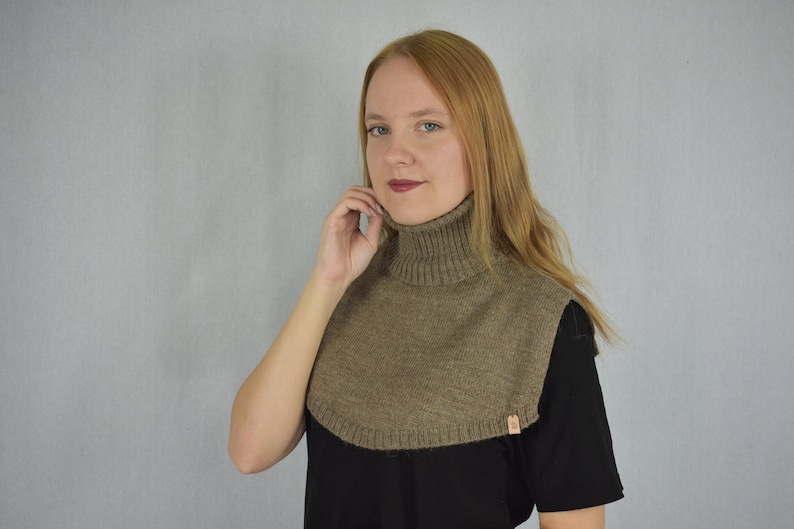 May include: A brown knitted turtleneck dickey featuring a ribbed collar and a rounded hemline that covers the upper chest and shoulders. The garment includes a small, rectangular tan brand tag attached to the lower edge of the knitted fabric.