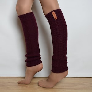 May include: A pair of dark burgundy knit leg warmers featuring a ribbed texture and a small rectangular tan leather tag with the text Hand Made attached to the upper side of the left leg warmer. The leg warmers extend from the ankle to the knee.
