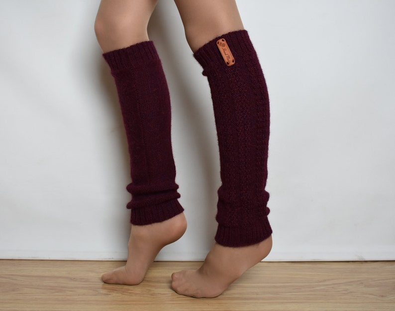 May include: A pair of dark burgundy knit leg warmers featuring a ribbed texture and a small rectangular tan leather tag with the text Hand Made attached to the upper side of the left leg warmer. The leg warmers extend from the ankle to the knee.