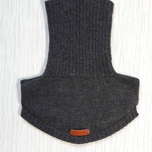 May include: A dark charcoal gray knitted neck warmer featuring a tall ribbed collar and a wider, smooth-knit base that covers the upper chest and shoulders. A small, rectangular brown leather tag with the text Hand Made is attached at the bottom.