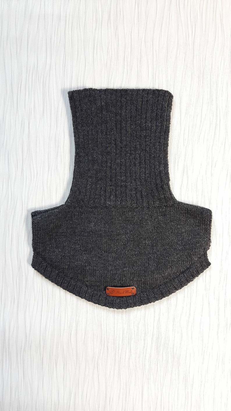 May include: A dark charcoal gray knitted neck warmer featuring a tall ribbed collar and a wider, smooth-knit base that covers the upper chest and shoulders. A small, rectangular brown leather tag with the text Hand Made is attached at the bottom.