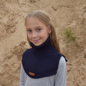 May include: A dark navy blue knitted neck warmer featuring a ribbed turtleneck collar and a smooth knit chest panel. The accessory includes a small rectangular brown leather-like brand tag attached to the lower front edge of the garment.