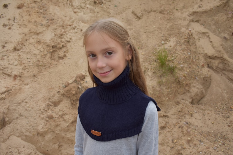 May include: A dark navy blue knitted neck warmer featuring a ribbed turtleneck collar and a smooth knit chest panel. The accessory includes a small rectangular brown leather-like brand tag attached to the lower front edge of the garment.