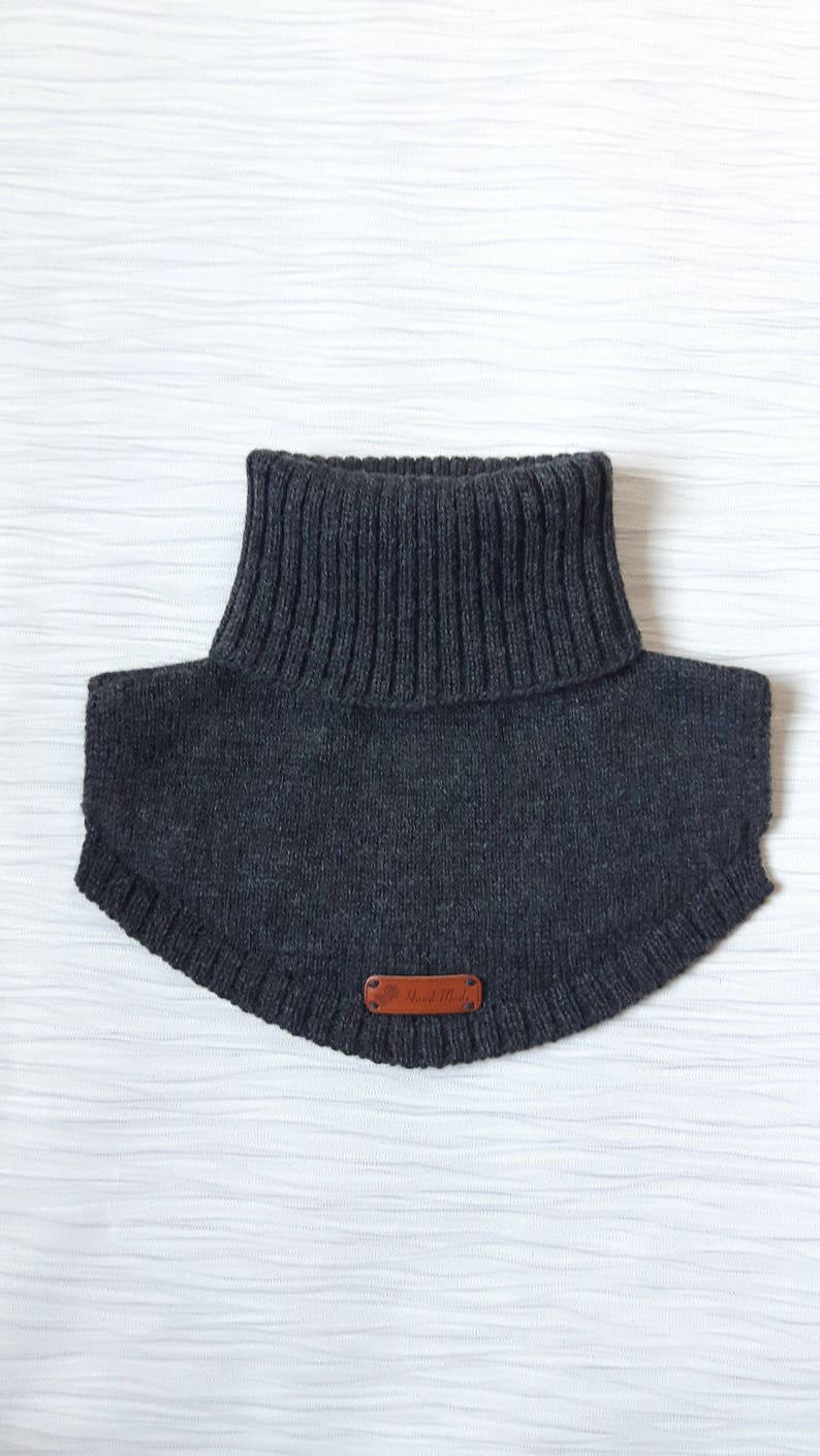 May include: A dark charcoal gray knitted neck warmer featuring a ribbed turtleneck collar and a curved hemline. A small, rectangular brown leather-like tag is stitched onto the lower front center, displaying the embossed text Hand Made.