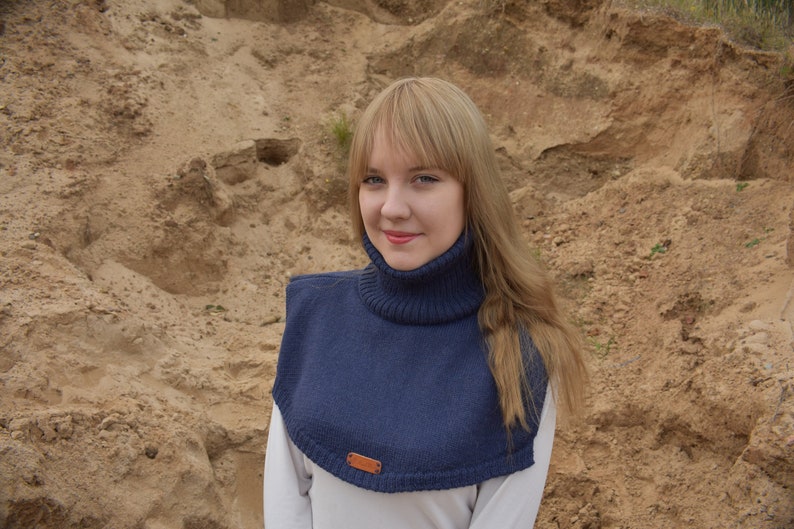 May include: A dark blue knitted cowl neck dickey featuring a ribbed turtleneck collar and a smooth knit chest panel. A small rectangular brown leather-like brand tag is attached to the bottom center edge of the garment, which is worn over a white shirt.