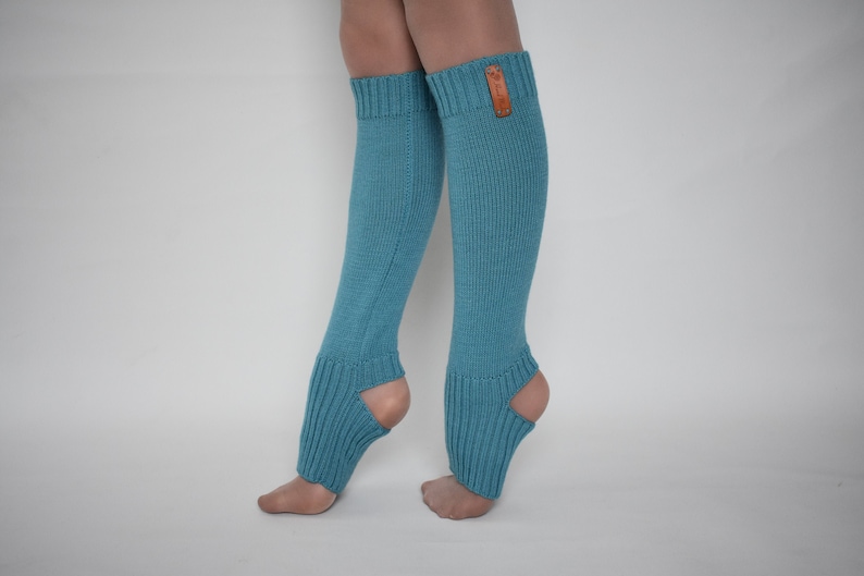 May include: A pair of teal blue knit leg warmers featuring a ribbed texture, an open heel design, and a small rectangular brown leather-like brand tag attached to the upper cuff of the right leg warmer, extending from the calf to the ankle.