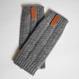 May include: A pair of grey knit fingerless gloves featuring a textured cable knit pattern, ribbed cuffs at both ends, and a rectangular tan leather tag attached with two silver-toned metal rivets near the top edge of each glove.