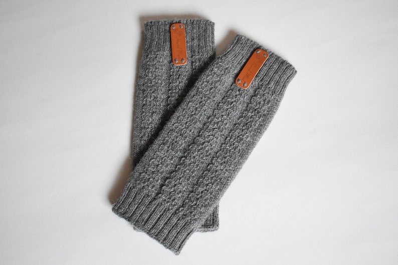 May include: A pair of grey knit fingerless gloves featuring a textured cable knit pattern, ribbed cuffs at both ends, and a rectangular tan leather tag attached with two silver-toned metal rivets near the top edge of each glove.