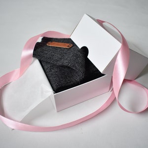 May include: A dark gray knitted garment folded inside a white cardboard gift box. The box is partially open and wrapped with a long, light pink satin ribbon. A small, rectangular brown leather-like tag is attached to the dark gray fabric.