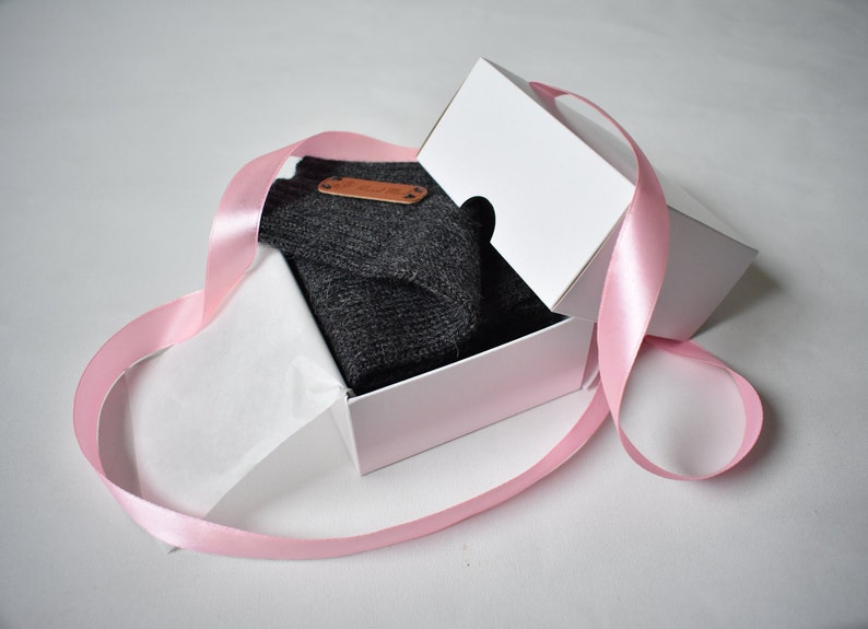 May include: A dark gray knitted garment folded inside a white cardboard gift box. The box is partially open and wrapped with a long, light pink satin ribbon. A small, rectangular brown leather-like tag is attached to the dark gray fabric.
