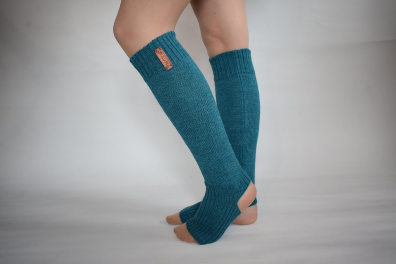 May include: A pair of teal, knee-high, open-heel leg warmers featuring a ribbed knit texture and a small, rectangular brown leather-like tag attached to the upper cuff. The design includes an opening for the heel and toes, exposing the skin.