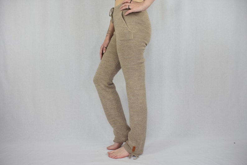 Puede incluir: Pantalones de punto beige con cintura de cord&oacute;n, bolsillos laterales y una peque&ntilde;a etiqueta rectangular marr&oacute;n cerca del dobladillo. Los pantalones tienen una silueta de pierna recta con lazos ajustables en la parte inferior de cada pierna.