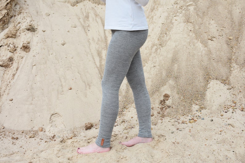 May include: A pair of heather gray leggings featuring a small brown rectangular leather-like patch near the ankle, paired with a plain white long-sleeved top. The leggings have a fitted silhouette and a textured knit appearance.