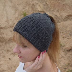 Peut inclure: Un bonnet en tricot gris avec un trou au sommet pour une queue de cheval. Le bonnet est porté par une personne aux cheveux blonds.