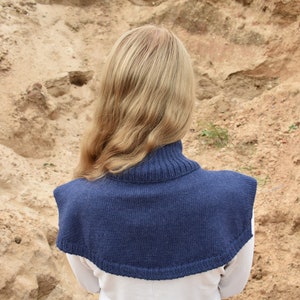 May include: A dark blue knitted cowl neck dickey featuring a ribbed collar and a wide, rounded shoulder covering. The garment is constructed from a solid-colored yarn with a consistent knit texture, providing coverage over the upper chest and back.
