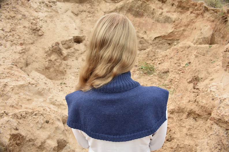 May include: A dark blue knitted cowl neck dickey featuring a ribbed collar and a wide, rounded shoulder covering. The garment is constructed from a solid-colored yarn with a consistent knit texture, providing coverage over the upper chest and back.