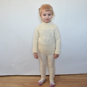 May include: A young child wearing a white knitted sweater and matching pants. The child is standing in front of a white wall.