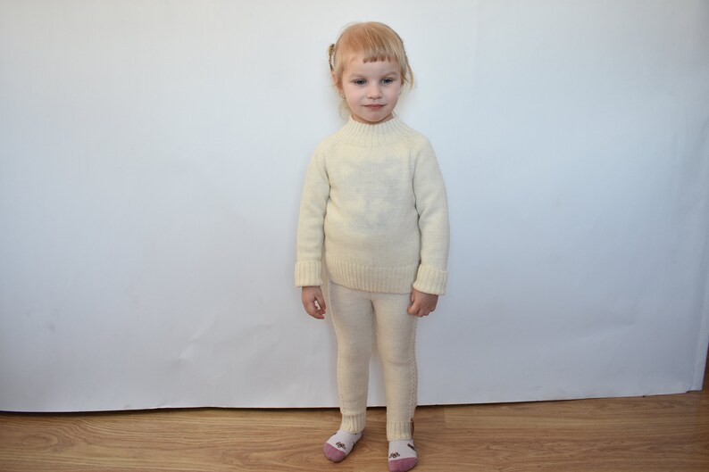 May include: A young child wearing a white knitted sweater and matching pants. The child is standing in front of a white wall.