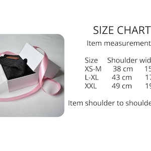 May include: A dark grey garment inside a white gift box wrapped with a pink ribbon. A size chart is displayed with the text: SIZE CHART, Item measurements, Size, Shoulder width, XS-M 38 cm 15", L-XL 43 cm 17", XXL 49 cm 19", Item shoulder to shoulder width.