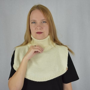 May include: A cream-colored knitted turtleneck dickey featuring a ribbed collar and a rounded hemline that covers the upper chest and shoulders. The garment is constructed with a smooth knit texture and a thick, folded ribbed neck section.