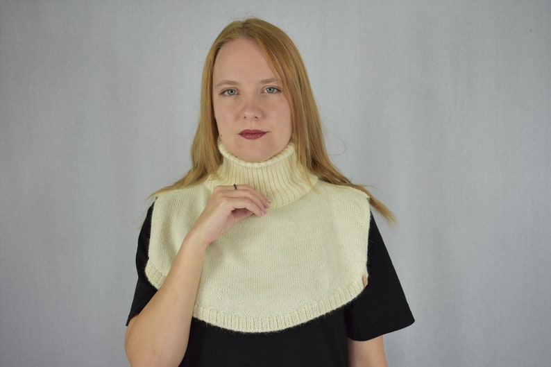 May include: A cream-colored knitted turtleneck dickey featuring a ribbed collar and a rounded hemline that covers the upper chest and shoulders. The garment is constructed with a smooth knit texture and a thick, folded ribbed neck section.