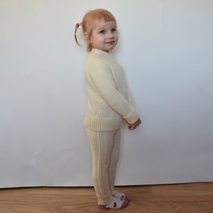 May include: A young child wearing a cream-colored knitted sweater and matching pants. The child has blonde hair and is standing in front of a white wall.