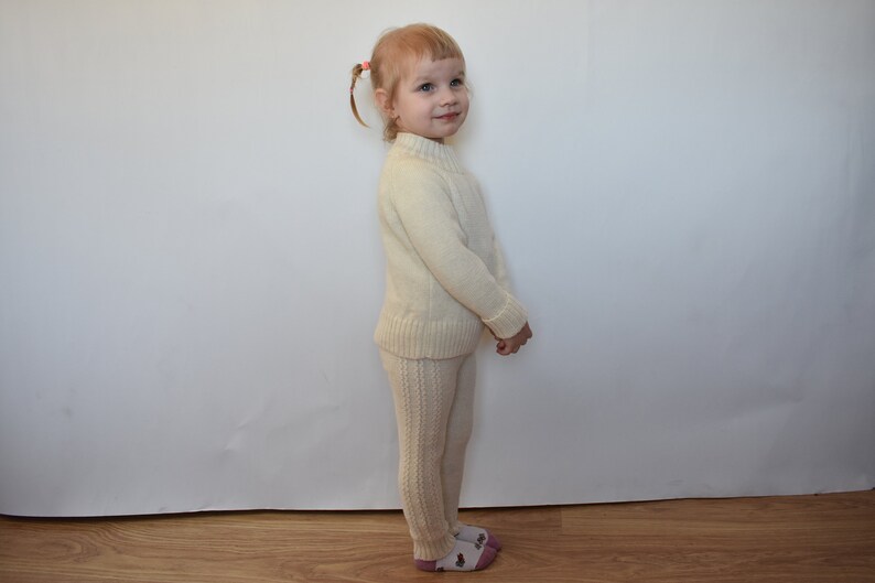 May include: A young child wearing a cream-colored knitted sweater and matching pants. The child has blonde hair and is standing in front of a white wall.
