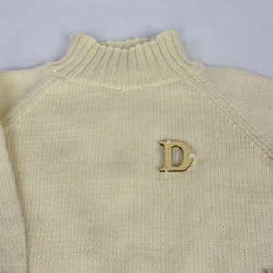 May include: A cream colored knitted sweater with a mock neck and a wooden letter 'D' pinned to the front.