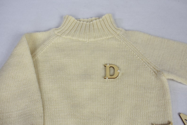 May include: A cream colored knitted sweater with a mock neck and a wooden letter 'D' pinned to the front.