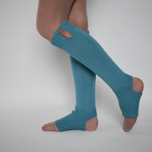 May include: A pair of teal blue knit leg warmers featuring a ribbed texture and an open heel design. The leg warmers extend from the mid-calf down to the foot, with a small rectangular brown leather-like tag attached to the upper ribbed cuff.
