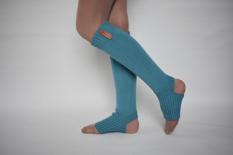 May include: A pair of teal blue knit leg warmers featuring a ribbed texture and an open heel design. The leg warmers extend from the mid-calf down to the foot, with a small rectangular brown leather-like tag attached to the upper ribbed cuff.