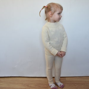 May include: A white knitted sweater and matching pants set for a child. The sweater has a mock neck and long sleeves. The pants are slim-fitting and have a straight leg.