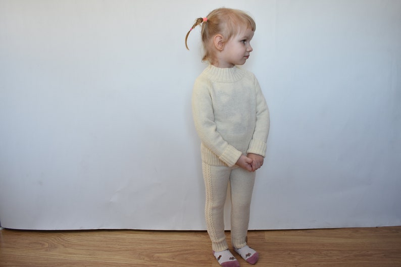 May include: A white knitted sweater and matching pants set for a child. The sweater has a mock neck and long sleeves. The pants are slim-fitting and have a straight leg.