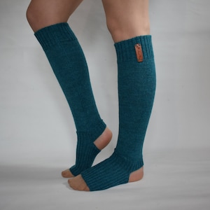 May include: A pair of teal, ribbed knit leg warmers featuring an open-heel design and a small, rectangular brown leather-like tag attached to the upper cuff. The knit fabric has a textured appearance and extends from the calf down to the mid-foot.