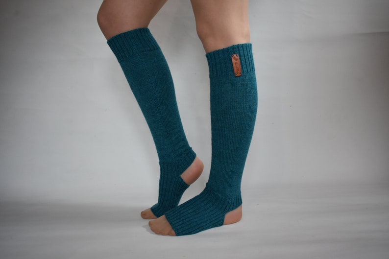 May include: A pair of teal, ribbed knit leg warmers featuring an open-heel design and a small, rectangular brown leather-like tag attached to the upper cuff. The knit fabric has a textured appearance and extends from the calf down to the mid-foot.