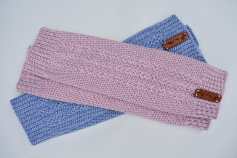 May include: A pair of knitted leg warmers in light pink and periwinkle blue. Each piece features a ribbed cuff at both ends and a central textured cable knit pattern. A small brown rectangular leather tag labeled Hand Made is attached.