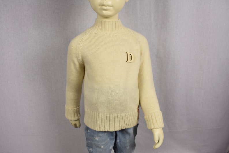 May include: A cream colored knitted sweater with a mock neck and a wooden letter 'D' on the front.