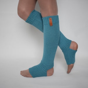 May include: Teal knit leg warmers featuring a ribbed texture, an open heel design, and a small rectangular brown leather tag attached to the upper cuff. These footless socks extend from the mid-calf down to the mid-foot, providing coverage.