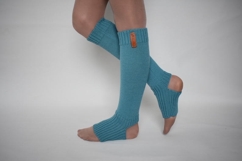 May include: Teal knit leg warmers featuring a ribbed texture, an open heel design, and a small rectangular brown leather tag attached to the upper cuff. These footless socks extend from the mid-calf down to the mid-foot, providing coverage.
