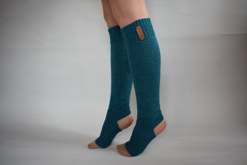 May include: A pair of teal, knee-high, ribbed knit leg warmers featuring an open heel and toe design. The leg warmers have a textured, knitted appearance and include a small, rectangular brown leather-like brand tag attached to the upper cuff.